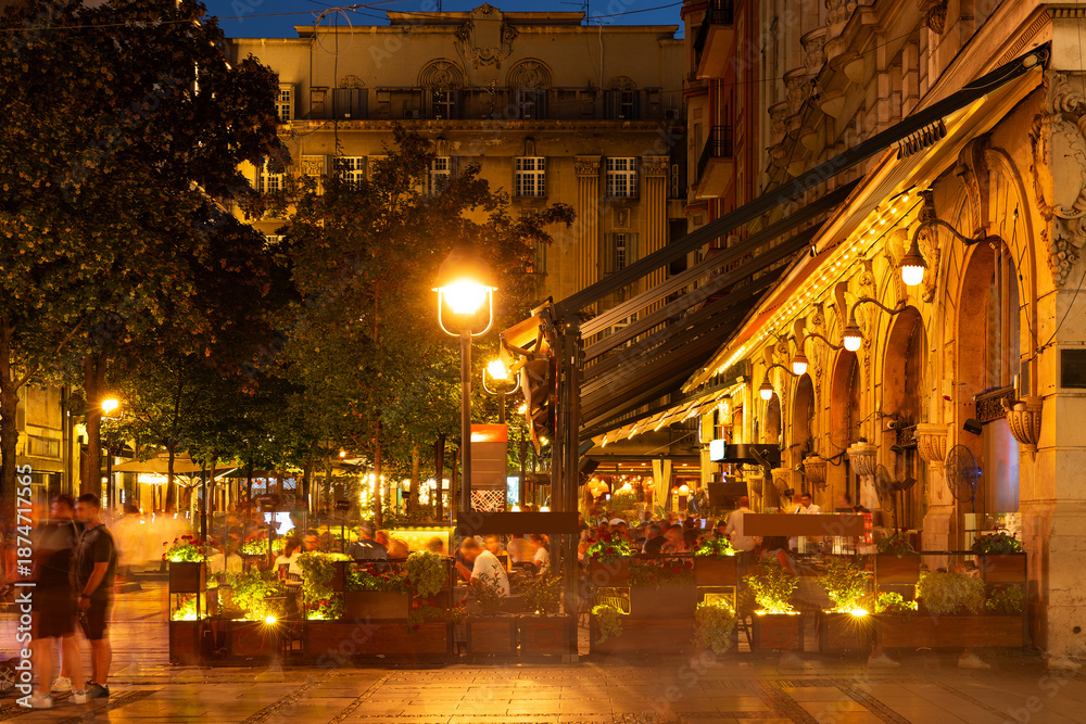Fototapeta premium Popular and bustling street cafe on Knez Mihailova Street in Belgrade after dark, with people dining outside under warm lights and canopy of trees on summer night