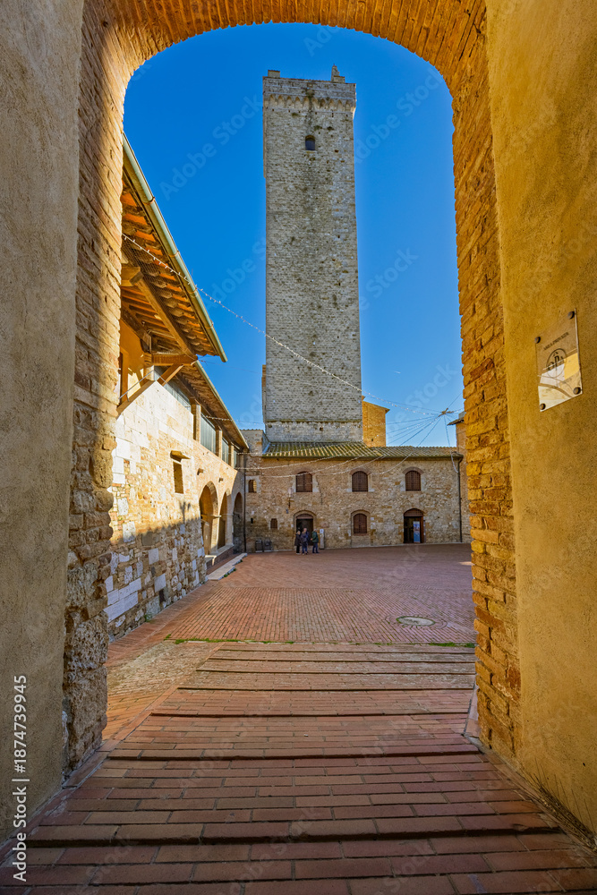 Fototapeta premium A view of the Torre Grossa, the town's tallest medieval tower, framed by a historic brick archway in the heart of San Gimignano, Tuscany.