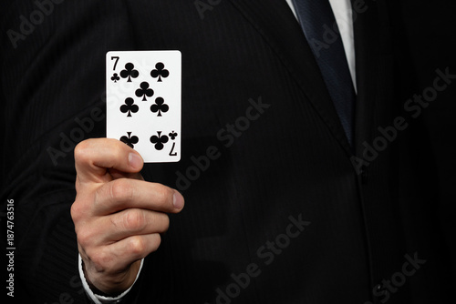 a businessman on suit holding a black seven of clovers card on his hand at the left side of the screen