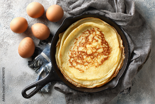 Stack of golden crepes in a cast iron pan with fresh eggs on the side © Tohamina