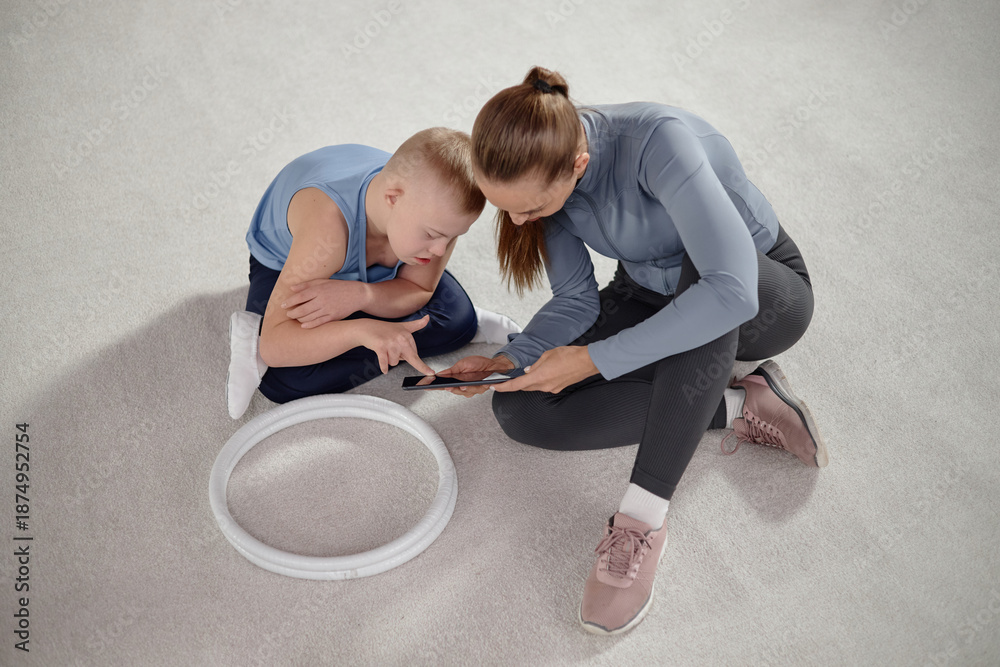 Naklejka premium Caucasian child with Down syndrome sitting on floor next to Caucasian woman, both interacting with digital tablet, child leaning in with interest, circular toy nearby