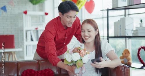Cheerful young Asian man surprises his girlfriend with a beautiful flower bouquet and a red gift box during a romantic celebration in a cozy, decorated living room.