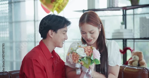 Cheerful young Asian man surprises his girlfriend with a beautiful flower bouquet and a red gift box during a romantic celebration in a cozy, decorated living room.