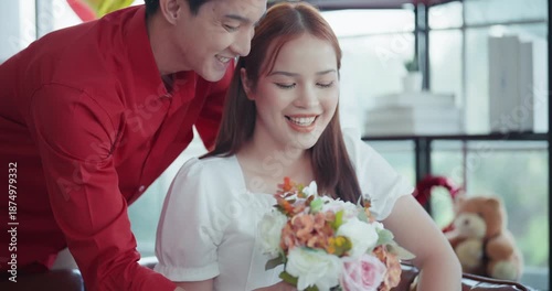 Happy young Asian man surprising his girlfriend with a beautiful flower bouquet and a red gift box, sharing a joyful and romantic moment together in a decorated room.