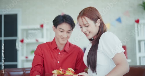 Smiling young Asian man and woman holding a red gift box with a gold ribbon, celebrating a special occasion or Valentine's Day in a decorated living room.