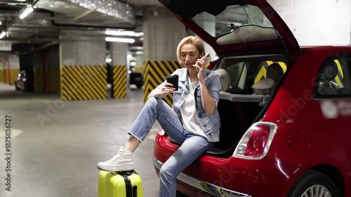 A woman sits in the open trunk of a red car in a parking garage while talking on her phone and holding a coffee cup