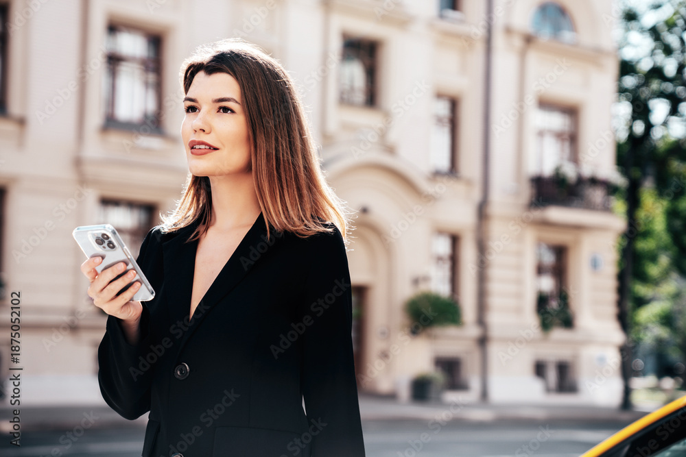 Fototapeta premium Young beautiful woman in nice trendy white long jacket suit. Sexy smiling model posing in street. Fashionable female outdoors. Cheerful and happy. Holds smartphone, uses phone apps, looks at screen