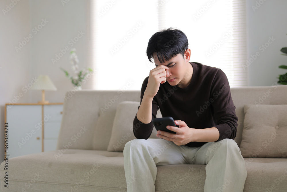 Obraz premium Young man sitting on a sofa, looking upset while holding a smartphone, experiencing headache and feeling disappointed