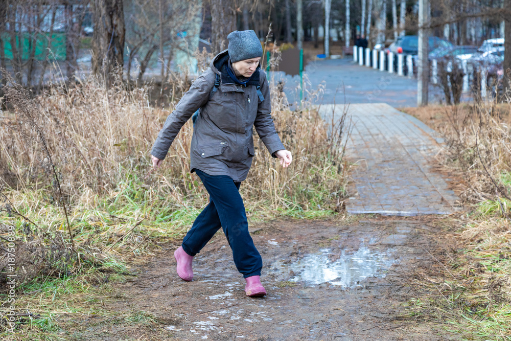 Fototapeta premium Caucasian woman walking on muddy path in rural area, wearing warm coat and hat. Adventure travel and outdoor activity concept for autumn hike.
