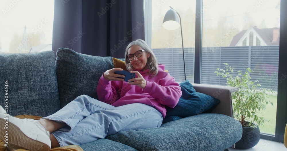 Fototapeta premium Elderly woman playing mobile phone lying on the sofa at home