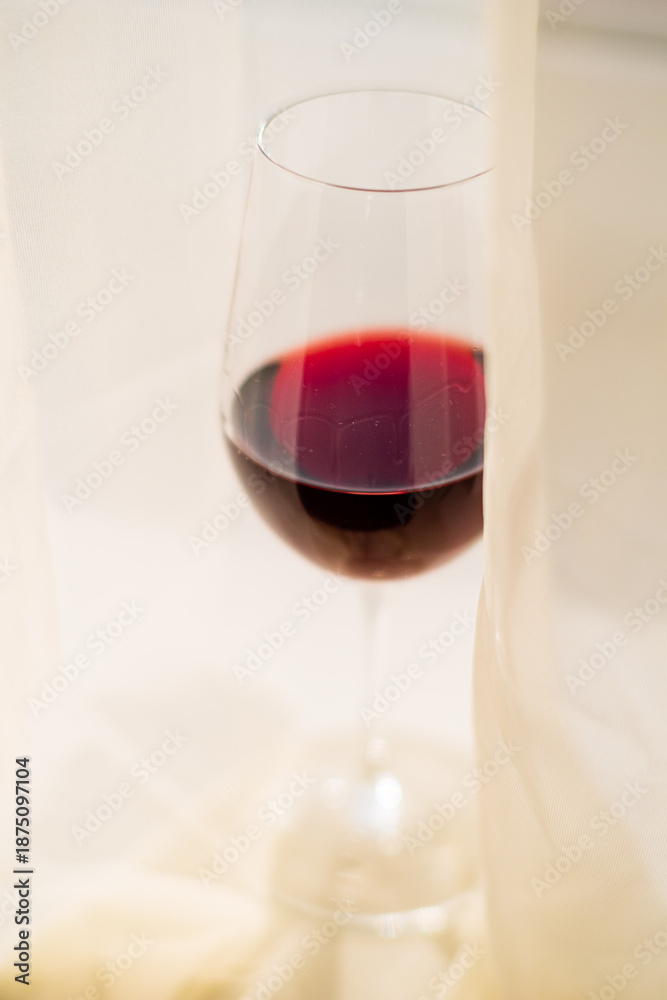 Fototapeta premium A wine glass filled with dark red liquid is partially visible behind soft, flowing curtains