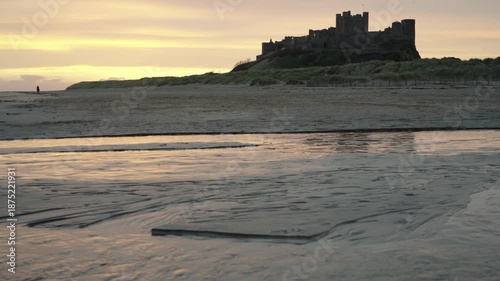 A low-angle static wide shot of the majestic Bamburgh Castle silhouetted against a golden sunrise sky, viewed from the sandy beach with a shallow stream reflecting the morning light in the foreground.