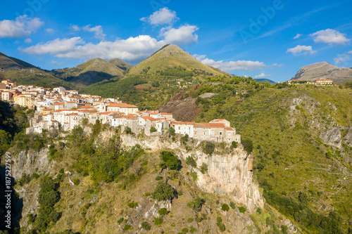 Fotografía Picturesque view of Tortora, Italy, with white stone houses and terracotta roofs perched atop a steep cliff, surrounded by lush green hills and rugged mountains under a bright blue sky
