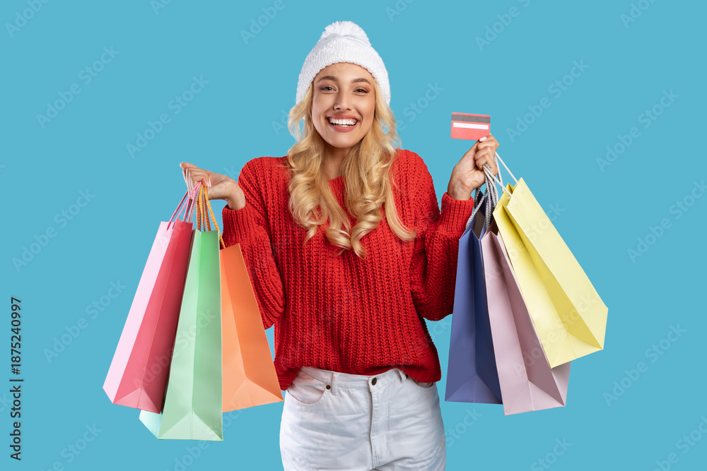 Obraz premium Winter Sale. Portrait of excited young lady in white hat holding credit card and colorful shopper bags from mall, buying gifts and presents using contactless payment, standing isolated at blue studio