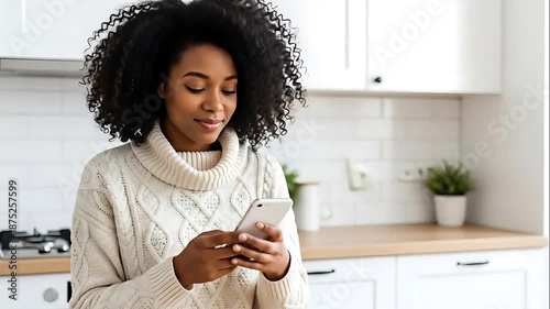 Woman in cozy sweater using smartphone in modern kitchen with white cabinets and plants