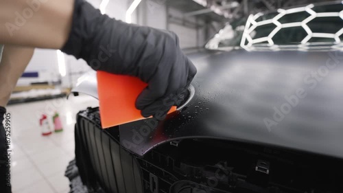 Automotive specialist carefully applying a protective paint film wrap on a modern black car using a squeegee tool in a detailing garage