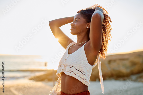 Quadro su tela Young black woman with curly hair by the ocean, her hands in her hair, looking o