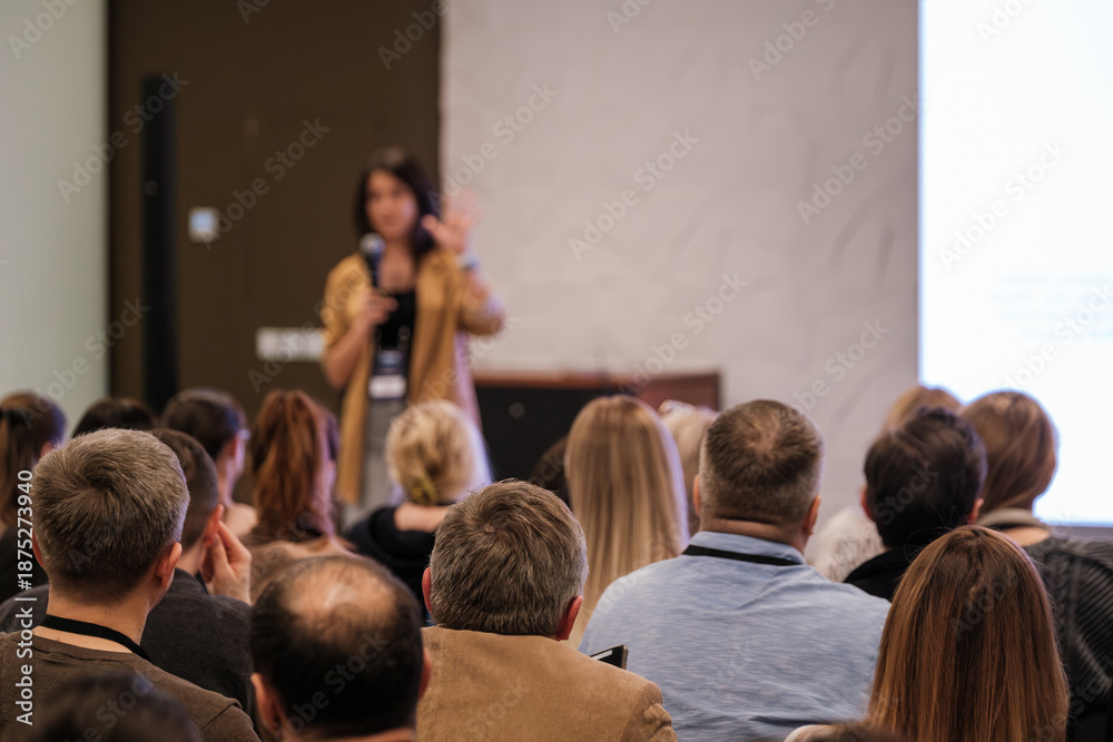 Obraz premium A confident presenter speaks to a seated audience in a conference room. Attendees listen attentively as a large screen glows in the background. Professional event atmosphere and learning environment.