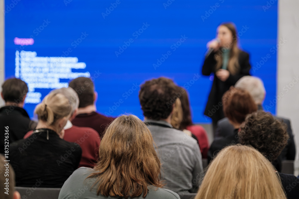 Obraz premium Audience seated facing speaker in conference room blue screen backdrop project slides professional presentation atmosphere.