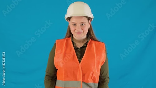Surprised young woman engineer takes paper bag with gift against blue wall. Curious lady engineer in helmet and reflective vest looks at present