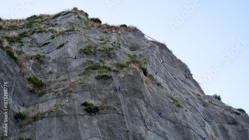 A low-angle detailed view of a steep rocky cliff face reinforced with a heavy-duty steel wire mesh netting designed to prevent rockfalls and landslides.