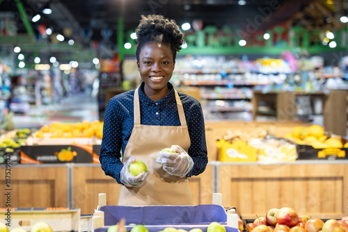 Billede på lærred African american woman employee smiling, wearing an apron and gloves, holding gr