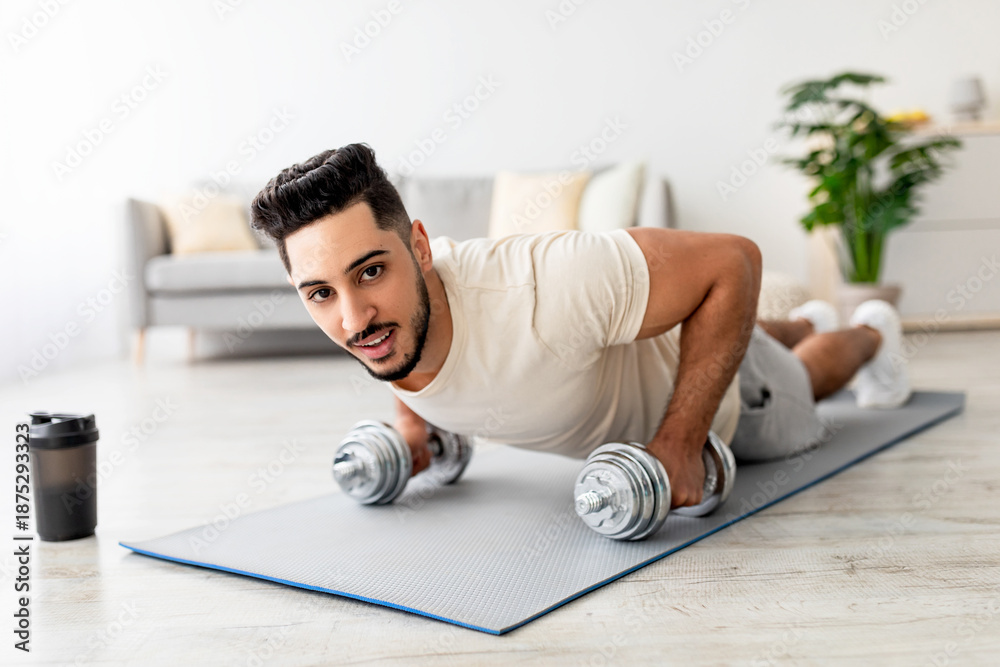 Fototapeta premium Stay at home fitness. Strong young Arab man doing whole body workout, standing in plank pose, using dumbbells indoors. Fit Eastern guy strengthening abs muscles in living room