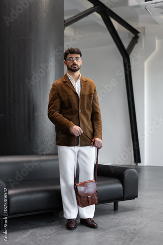 Stylish man in a brown corduroy blazer and white trousers holding a cognac leather messenger bag in a modern minimalist studio interior.