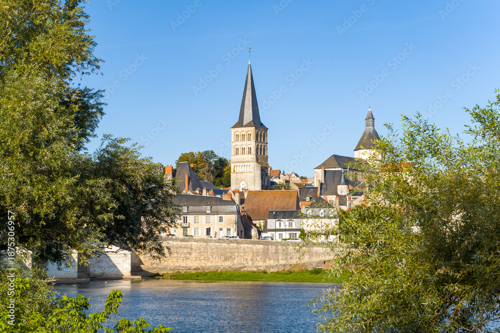 Fototapeta premium Distinctive abbey tower rises above the historic rooftops of La Charite sur Loire, framed by leafy trees and the calm Loire river under a clear blue sky. The scene highlights the medieval architecture