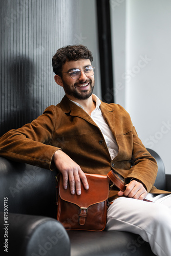 Sophisticated man in a brown corduroy blazer and glasses sitting on a black leather sofa holding a cognac leather messenger bag in a minimalist studio.