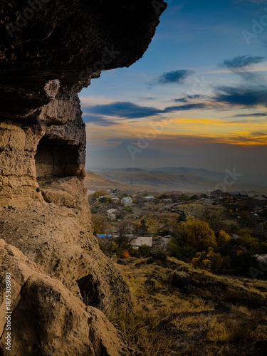 A rugged cave opening frames a picturesque landscape of a distant town and rolling hills under a clear sky. Sunlight highlights the warm tones of the rock formations surrounding the entrance. High
