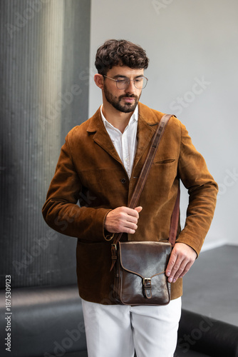 Portrait of a stylish man in a brown corduroy blazer and white shirt holding a cognac leather messenger bag in a minimalist studio.