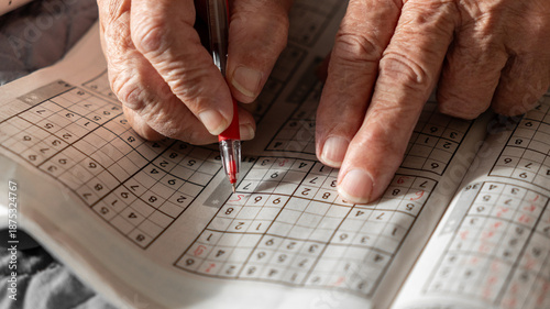 Photos old woman, pensioner, grandmother solves Sudoku crosswords and writes numbers in a journal with a red pen