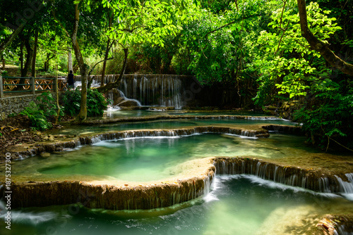 Sunlight filters through lush green foliage onto tiered turquoise pools and limestone cascades at Kuang Si Falls in northern Laos. The tranquil scene features clear water, natural terraces, and dense