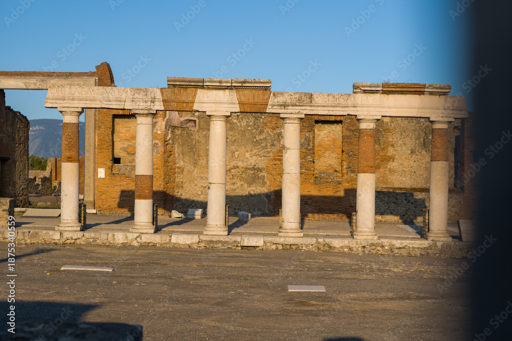Fototapeta premium Ancient marble columns and brick entablature are illuminated by warm evening light in the civil forum of Pompeii, Italy. The textured stonework and faded inscriptions stand out against the open plaza