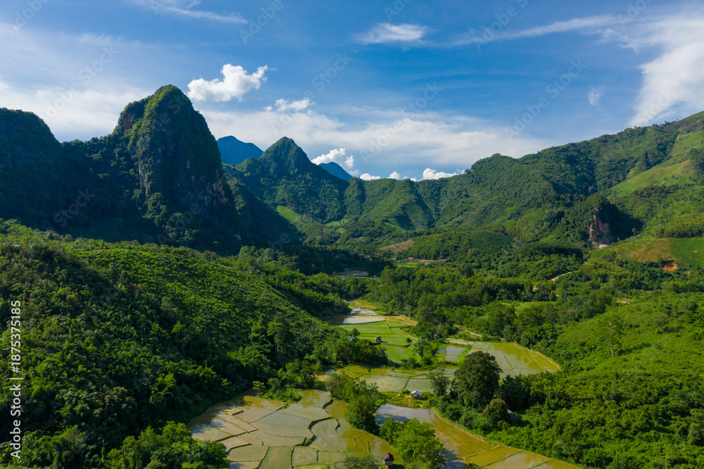 Fototapeta premium Aerial view of flooded rice paddies set within a green valley surrounded by steep limestone mountains and dense tropical forest in northern Laos. Bright daylight and blue sky highlight the layered