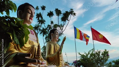 Two serene sitting Buddha statues with colorful Buddhist flags at Chua Da Bao Pagoda in Nha Trang, Vietnam. Peaceful spiritual scene in a traditional temple surrounded by nature and mountain views.