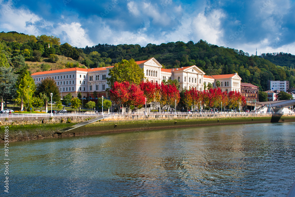 Obraz premium The modern skyline and historic industrial cranes mirrored in the calm, winding waters of the Bilbao river estuary.