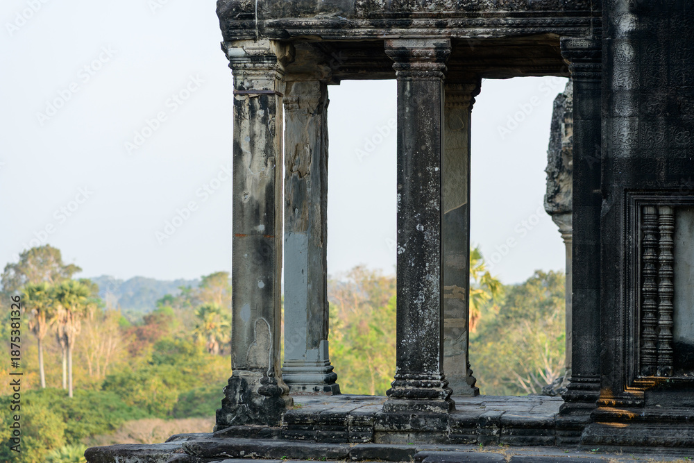 Fototapeta premium Aged sandstone pillars frame the view from the edge of Angkor Vat, Cambodia, revealing tropical foliage and soft morning light beyond. The textured surfaces and subtle carvings highlight the temple's