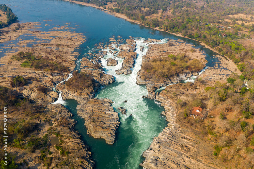Obraz premium Expansive aerial scene of the Chutes de Khone waterfalls on the Mekong River in southern Laos. Whitewater rapids cascade through rugged rocky channels, surrounded by dry forest and a wide river plain