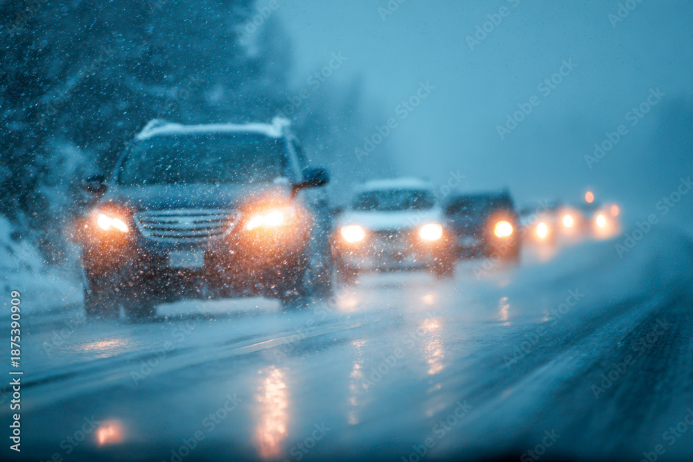 Obraz premium A line of vehicles driving cautiously on a slippery snow-covered highway with bright headlights glowing through heavy snowfall and foggy winter conditions