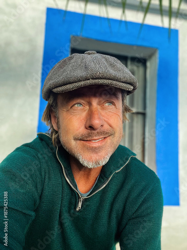 Tableau sur toile Smiling rugged man with woolen newsboy cap and stubble beard in green woolen sweater outside by white wall with window of spanish house