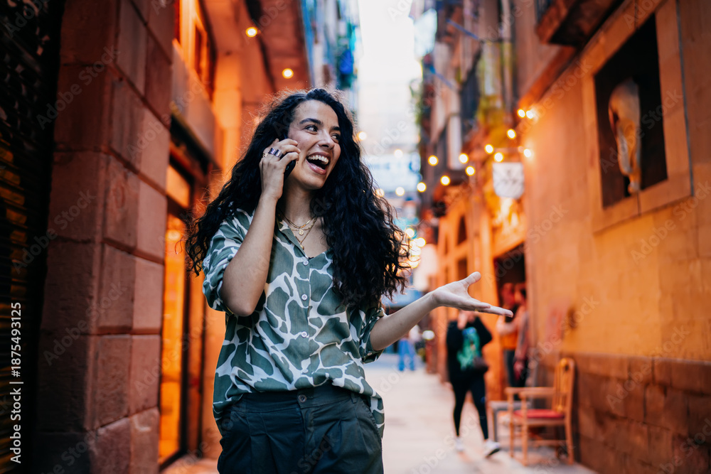 Obraz premium Excited woman gestures mid-call with expressive face, talking via smartphone in city alley, conveying engagement, connection and digital immersion in modern lifestyle.