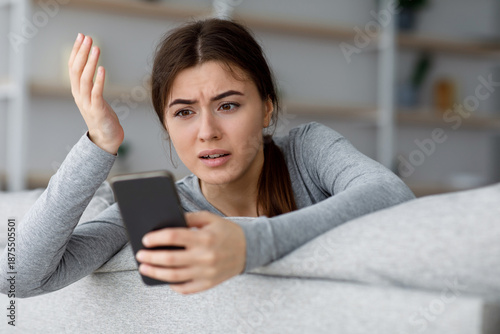 Fotografi Angry sad shocked depressive attractive young caucasian female looking at phone and gesturing, got bad news at home interior, free space