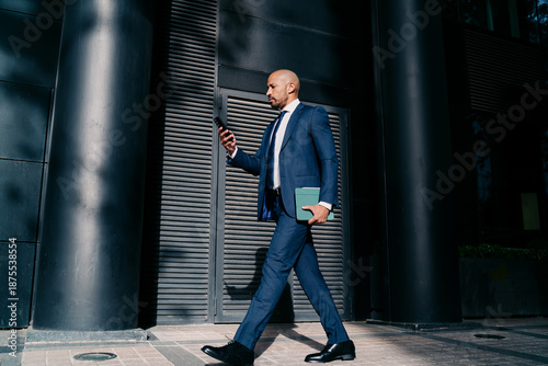 Canvas-taulu Focused businessman in tailored suit walking confidently in urban business district, multitasking with smartphone and digital tablet, symbolizing mobile decision-making and digital leadership
