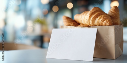 blank postcard mockup (thick cardstock) leaning against a simple white paper bag filled with croissants. The background is a soft blur of a bright, modern cafe interior