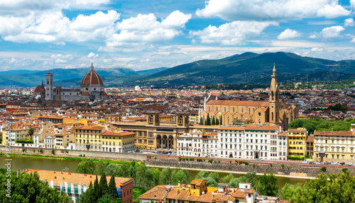 Panoramic view of Florence (Firenze), Italy, showing rooftops, landmarks like Palazzo Vecchio and Santa Maria del Fiore with its red dome, and hills with forests on a sunny summer day.
