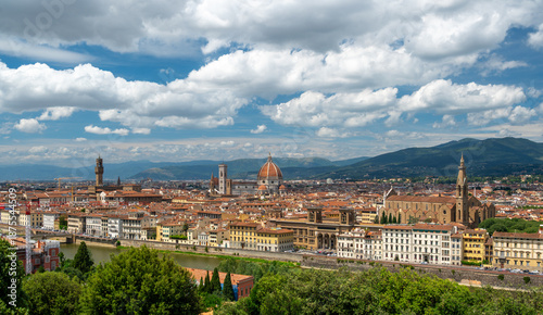Panoramic view of Florence (Firenze), Italy, showing rooftops, landmarks like Palazzo Vecchio and Santa Maria del Fiore with its red dome, and hills with forests on a sunny summer day.