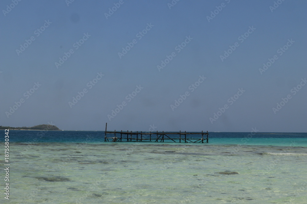 Fototapeta premium A tropical beach features a wooden pier stretching over turquoise ocean waves toward a blue horizon under a summer sky in this paradise island vacation landscape