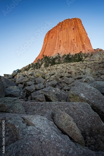 Devil's Tower glows orange in the evening light, rising above rocky slopes and pine trees in northeastern Wyoming.
